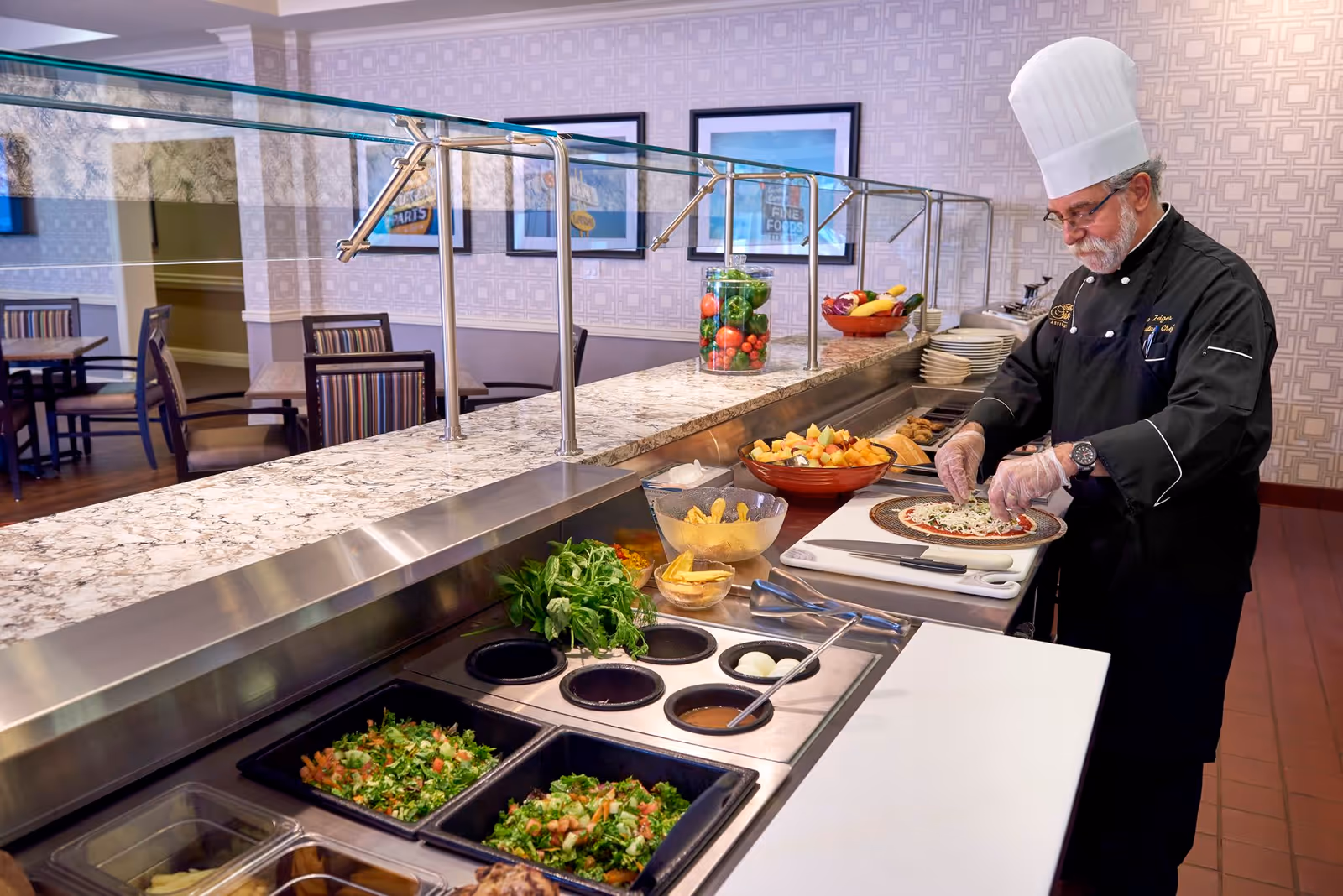 A chef wearing a white hat and black uniform prepares a pizza behind a buffet counter with various fresh ingredients and salads in a dining area with tables and chairs in the background.