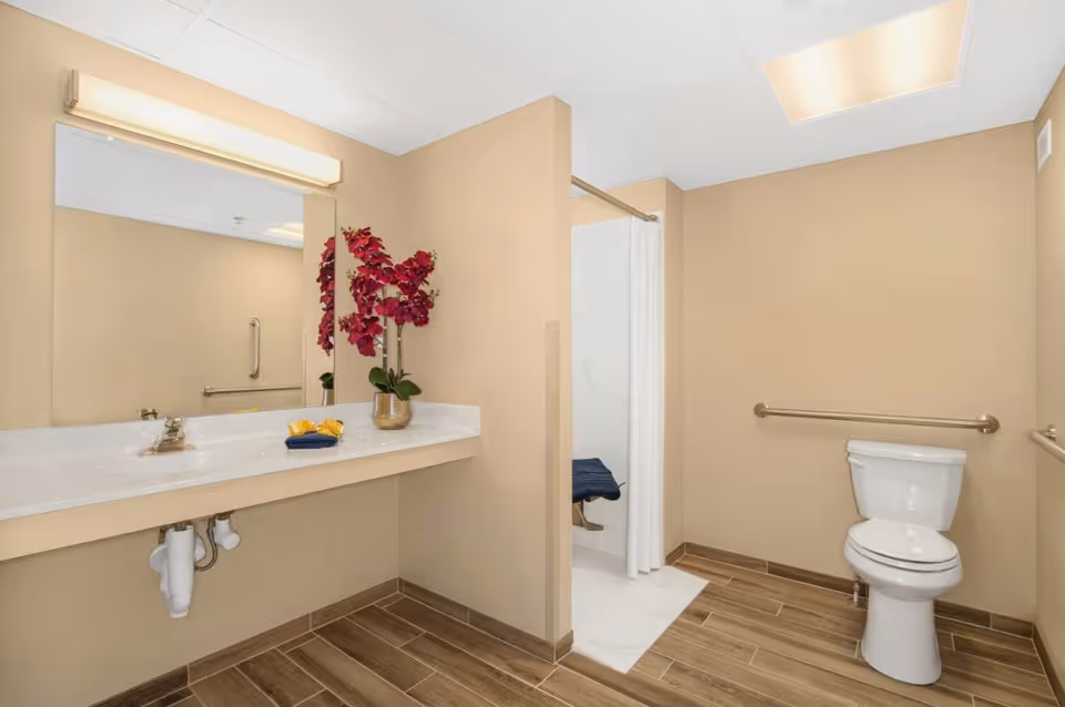 A clean and accessible bathroom with beige walls and wood-look tile flooring. The room features a white toilet with grab bars on the walls, a walk-in shower with a white curtain, and a long white countertop with a sink. On the countertop, there is a potted plant with red flowers and neatly folded towels. A large mirror with a horizontal light fixture above it is mounted on the wall above the sink.