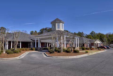 Exterior view of Dalton Square facility showing a single-story brick building with a covered entrance supported by white columns, a cupola on the roof, landscaped bushes and trees, and a clear blue sky.