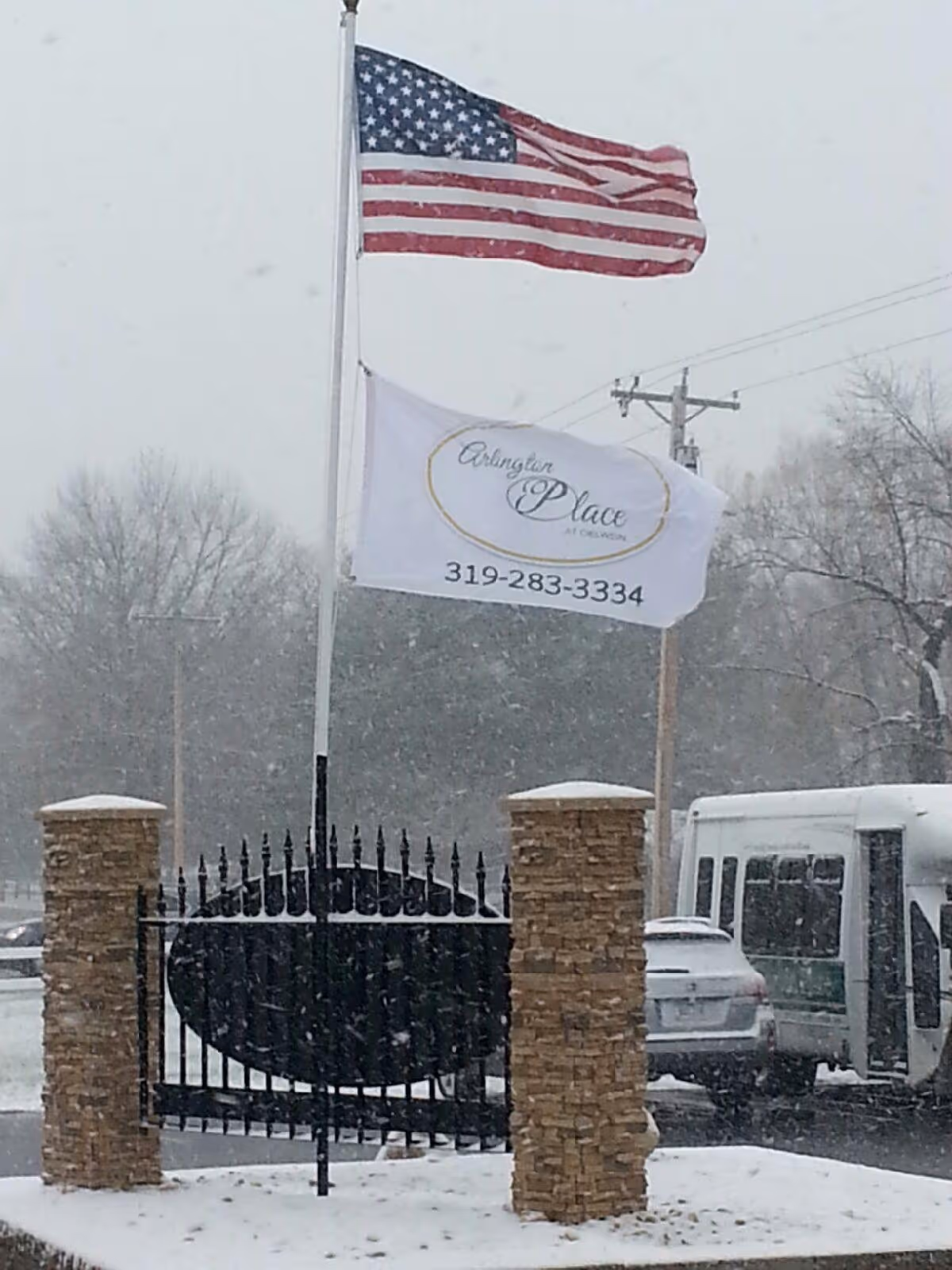 Two flags flying on flagpoles in a snowy outdoor setting. The top flag is the American flag, and the bottom flag is white with the text 'Arlington Place at Oelwein' and a phone number 319-283-3334. The flags are positioned behind a black metal gate with stone pillars on either side. There are trees and vehicles visible in the background.