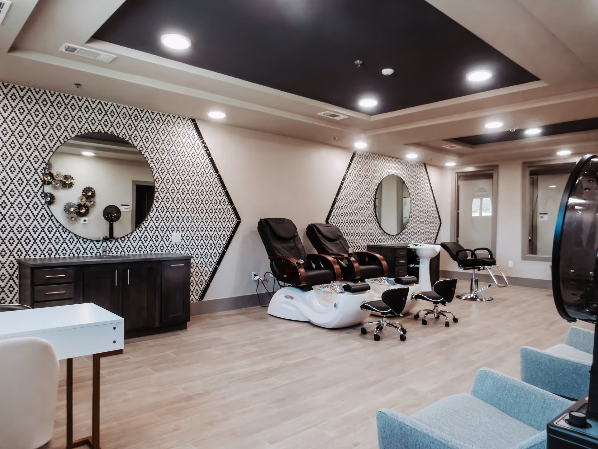 Interior of a salon area in a senior living facility with two black pedicure chairs, two round mirrors on patterned walls, a white manicure table, and light blue armchairs. The room has wooden flooring and recessed ceiling lights.