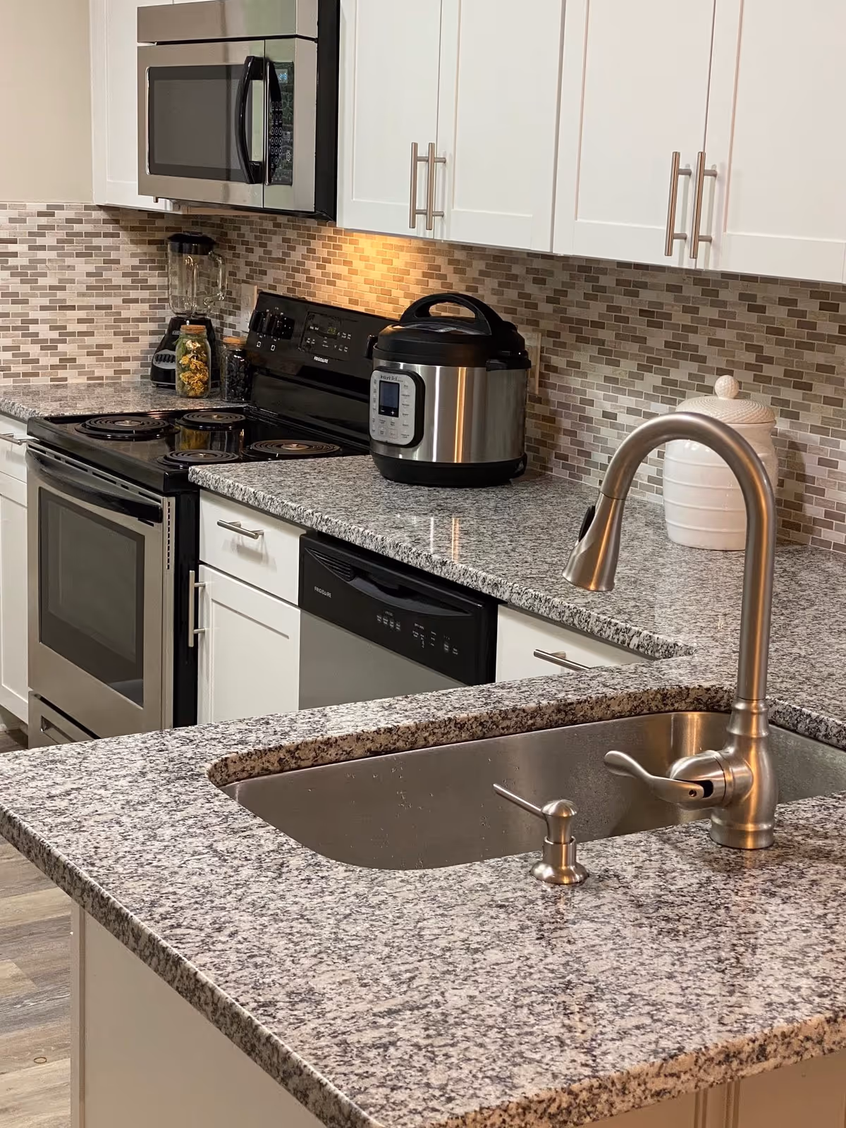Modern kitchen with granite countertops, a stainless steel sink with a faucet, a black electric stove, a microwave, a blender, an Instant Pot, and a white ceramic container. The backsplash features small rectangular tiles in neutral tones.