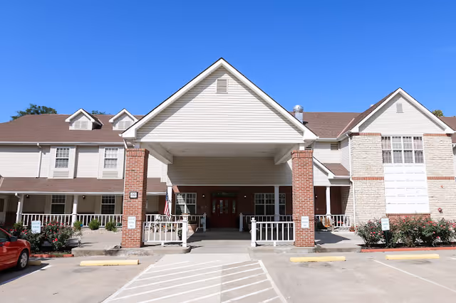 Front entrance of a senior living facility with a covered porte-cochere, columns, and adjacent parking area.