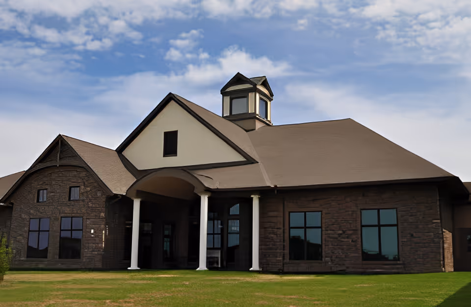 Exterior view of a large building with a brown roof, stone walls, multiple windows, and a small tower-like structure on top, set against a partly cloudy blue sky with green grass in the foreground.