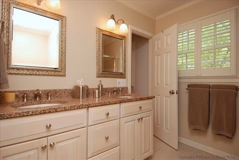 A bathroom with a double sink vanity featuring a granite countertop, two framed mirrors above each sink, and two wall-mounted light fixtures. There is a partially open door next to a window with white shutters, and two brown towels hanging on a towel rack beneath the window.