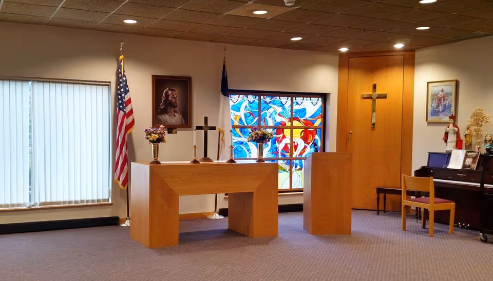Interior of a chapel or worship room with a wooden altar and podium, decorated with flowers, a cross, and religious statues. There is a stained glass window with colorful designs, an American flag, and a portrait of Jesus on the wall. A piano and chair are also visible on the right side.
