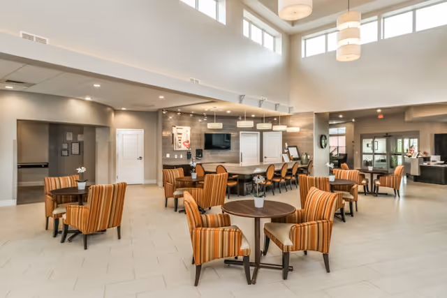 A spacious and well-lit senior living community common area with high ceilings and large windows. The room features multiple round wooden tables surrounded by striped upholstered chairs in warm tones. In the background, there is a long table with additional chairs, pendant lighting, a wall-mounted TV, and doors leading to other rooms. The area has a modern and welcoming atmosphere with light-colored tile flooring and neutral wall colors.