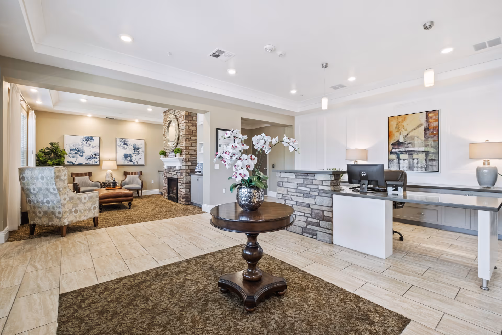 A bright and welcoming senior living facility lobby with a round wooden table holding a vase of white orchids in the center. To the right is a reception desk with a computer and two table lamps, and behind it is a colorful abstract painting. To the left is a cozy seating area with two armchairs, a leather ottoman, a stone fireplace with a round mirror above it, and two floral paintings on the wall. The floor is a combination of beige tiles and brown patterned carpet.