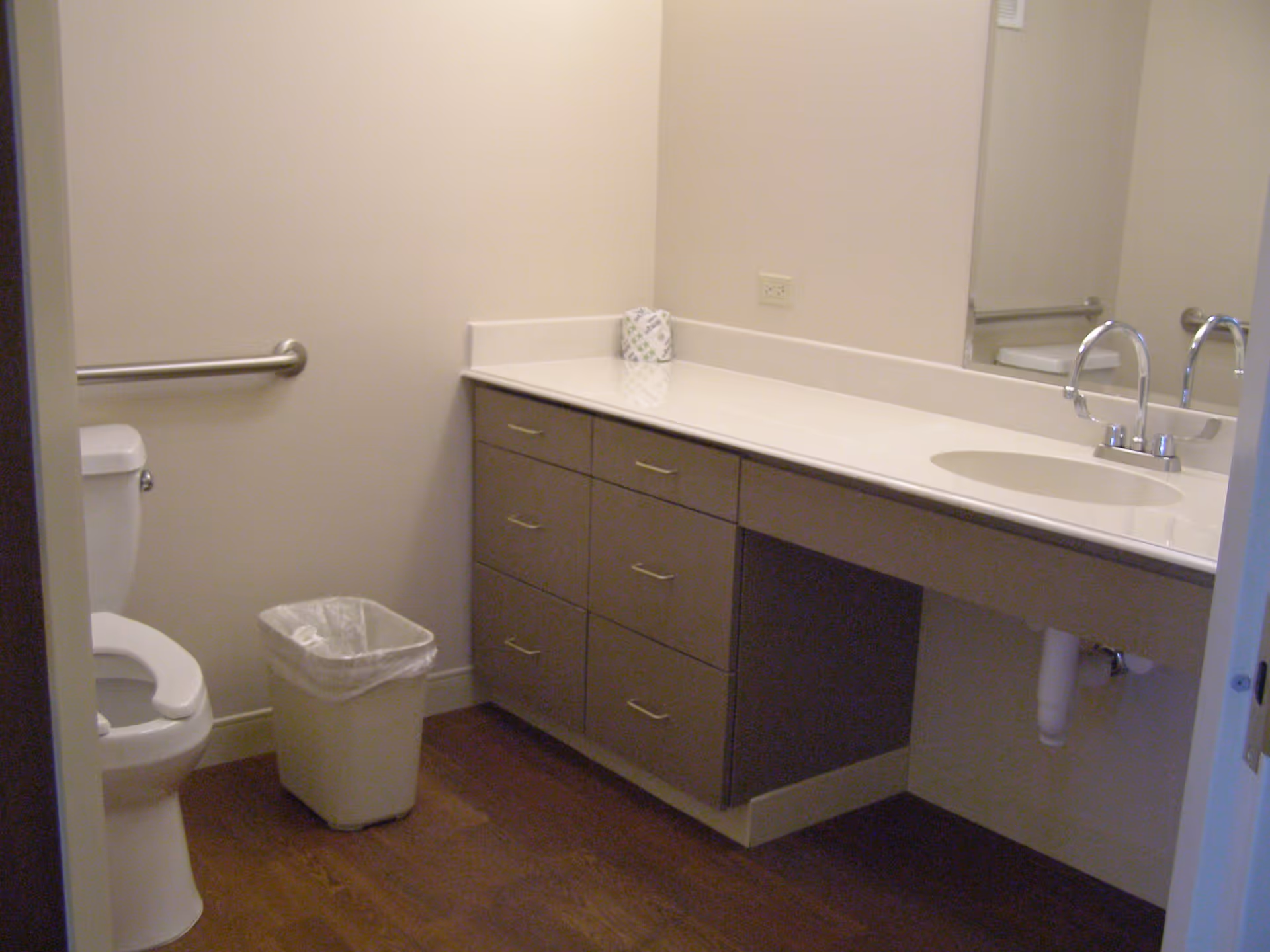 A clean bathroom with a white toilet, a trash can, a long countertop with a sink, multiple drawers, a large mirror, and a grab bar on the wall next to the toilet.