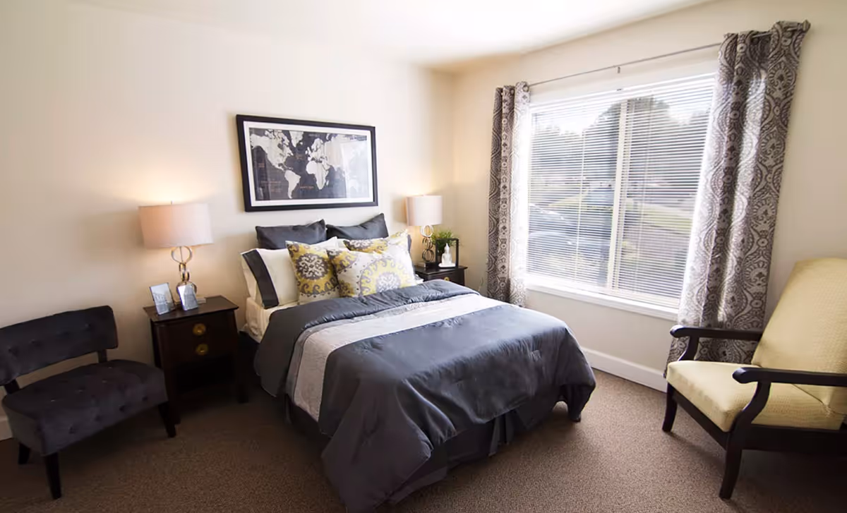 A cozy bedroom with a neatly made bed featuring gray and white bedding and decorative pillows. There are two bedside tables with lamps on each side of the bed, a framed world map hanging above the bed, a gray upholstered chair to the left, and a yellow armchair to the right near a large window with patterned curtains.