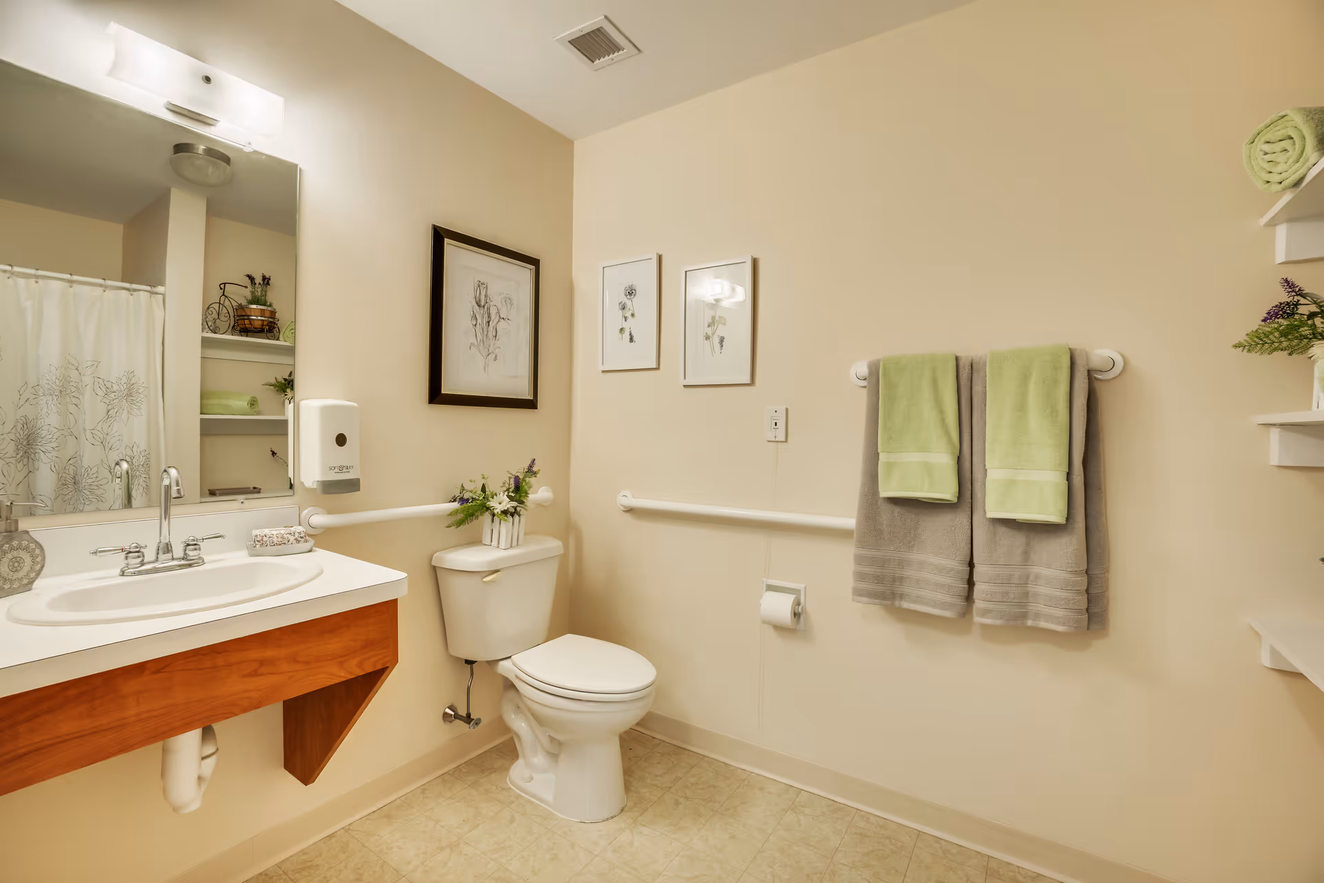 A clean and well-lit bathroom with a white toilet, a sink with a wooden vanity, a large mirror above the sink, and a shower curtain with a floral pattern. The walls are beige and decorated with framed botanical prints. Two sets of towels, one green and one gray, hang on a towel rack. There are grab bars near the toilet and sink for accessibility, and shelves with rolled towels and decorative plants.