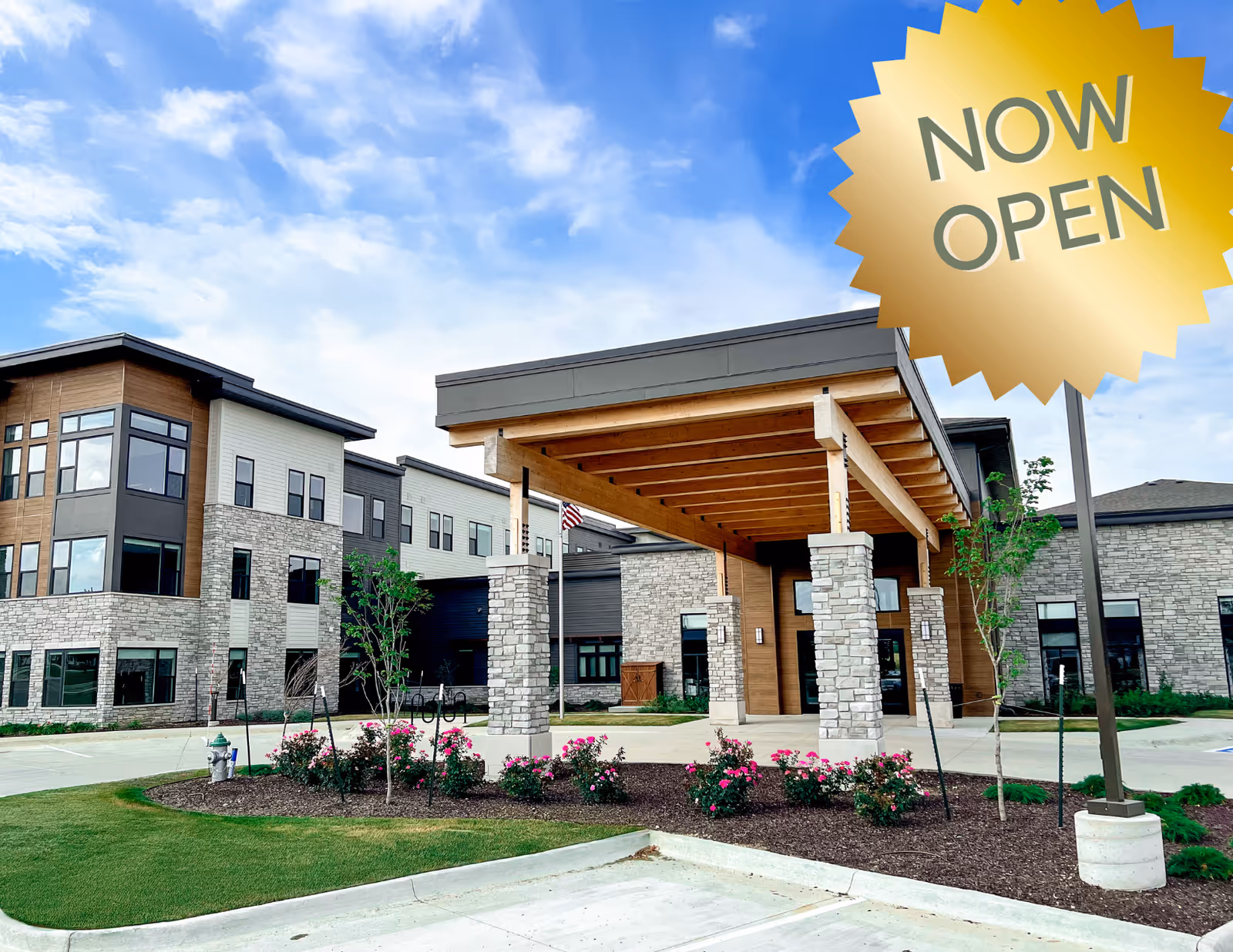 Front entrance of PrairieStone Senior Living showing the building exterior with a covered wooden porte-cochere, stone columns, and landscaped flower beds, with a "NOW OPEN" badge in the corner.