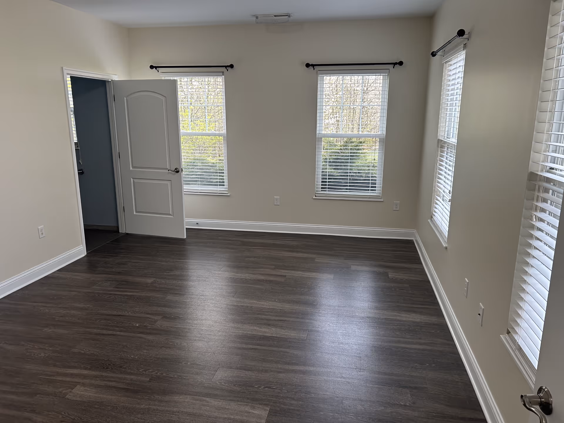 Empty room with dark wood flooring, beige walls, three windows with white blinds, and a white door partially open revealing a blue-painted adjacent room.