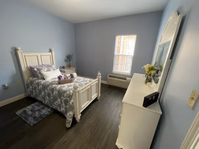 A small bedroom with light blue walls and dark wood flooring. The room features a white wooden bed with a floral gray and white bedspread, a small bedside table with a plant and decorative items, a white dresser with a large mirror, and a window letting in natural light. There is also a small rug beside the bed.