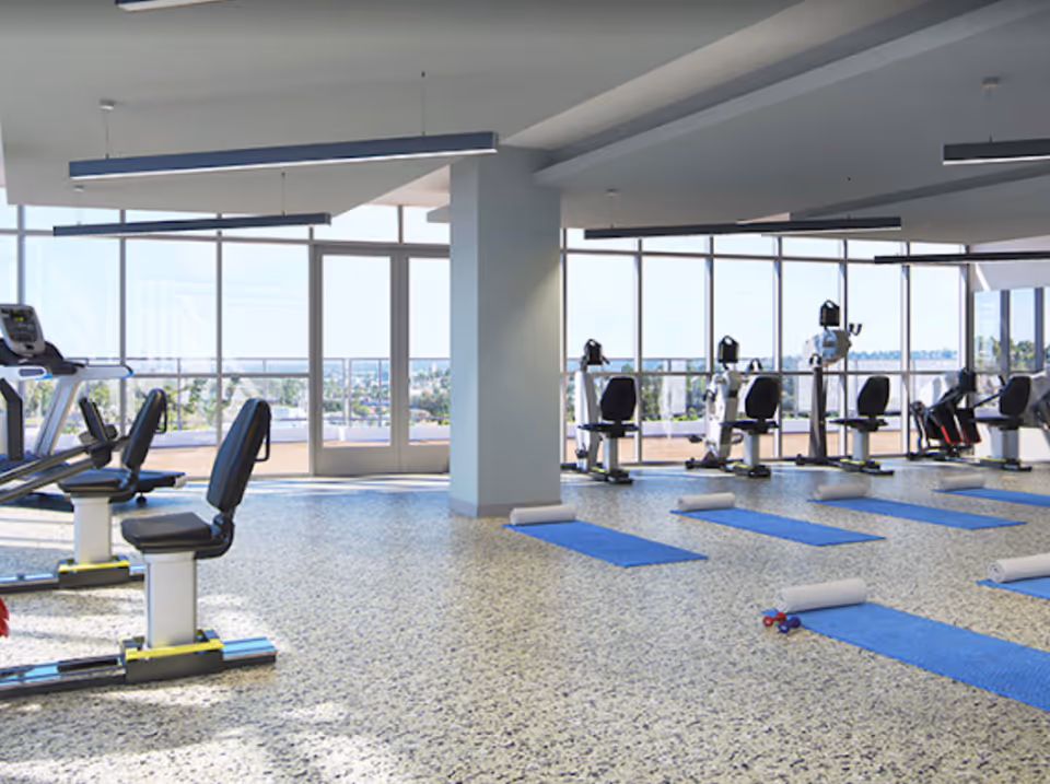 Bright fitness room with exercise machines and blue yoga mats arranged on a speckled floor facing large floor-to-ceiling windows.