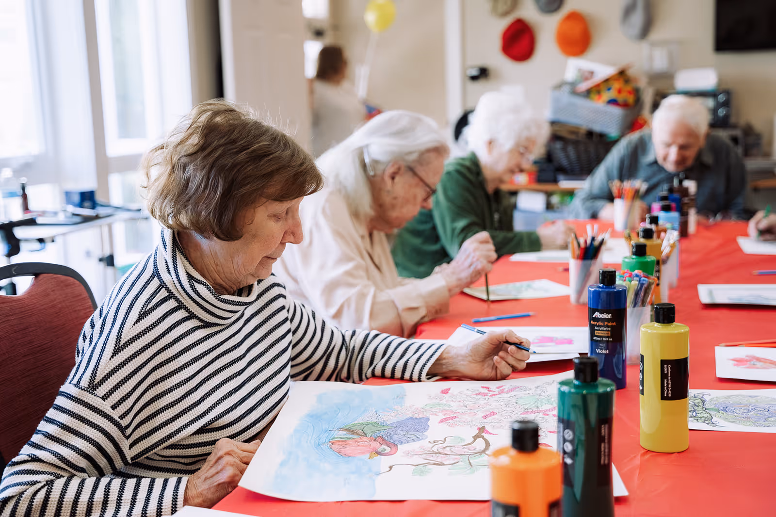 A group of elderly women sitting around a table covered with a red tablecloth, engaged in painting and coloring activities with various bottles of acrylic paint and colored pencils on the table in a well-lit room.