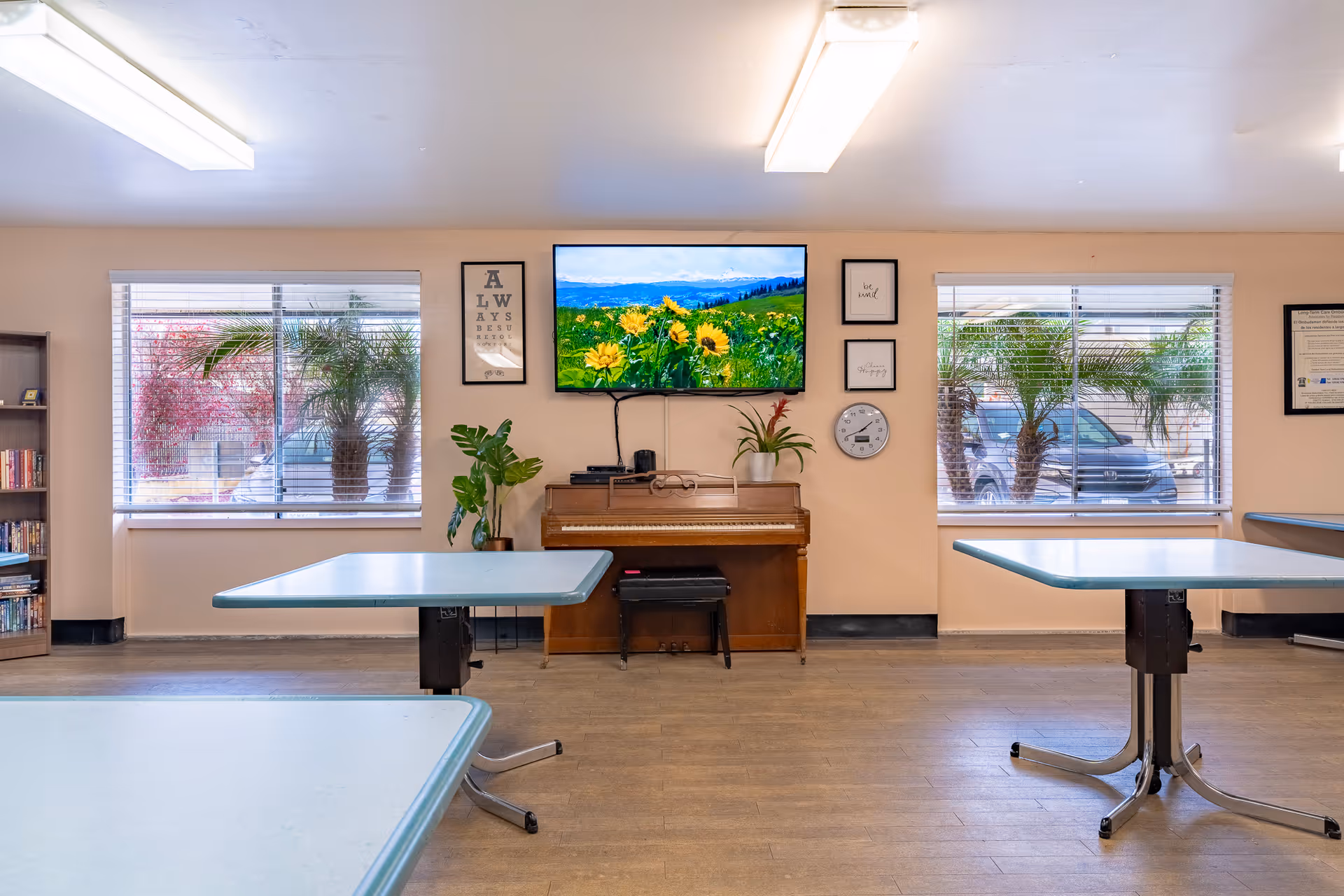 Community activity room with tables, a piano under a wall-mounted TV, plants, and windows with blinds.