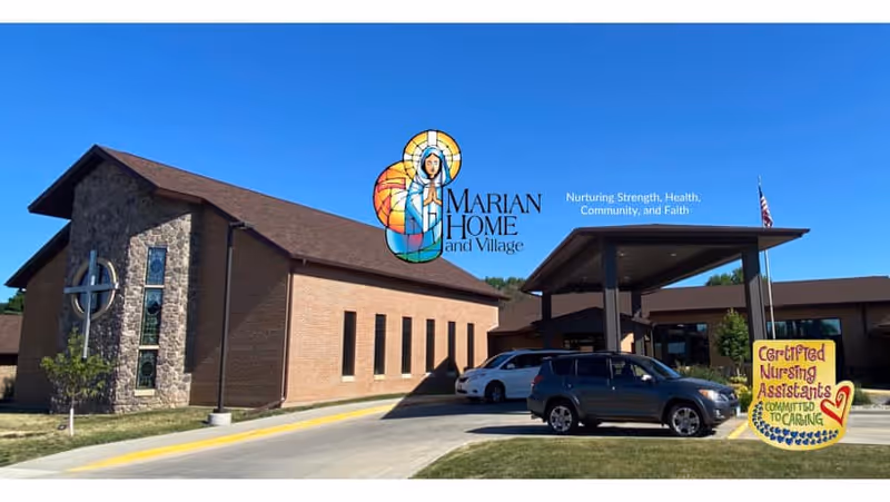 Exterior view of Marian Home and Village building under a clear blue sky. The building features a stone and brick facade with a large cross on the left side. Several cars are parked near the entrance, which has a covered drop-off area. A colorful sign near the entrance reads 'Certified Nursing Assistants Committed to Caring'.