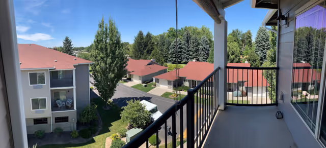 View from a second-floor balcony overlooking a senior living complex with red-roofed buildings, trees, and a driveway.