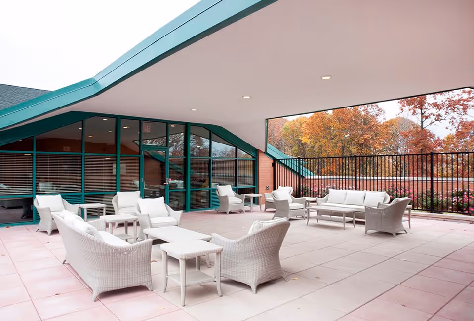 Outdoor covered patio area with white wicker seating including sofas, armchairs, and tables. The patio is adjacent to a building with large green-framed windows and doors. Trees with autumn foliage are visible beyond a black metal fence.