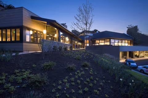 A modern senior living building at dusk with illuminated windows, a landscaped slope in the foreground, and parked cars by the entrance.
