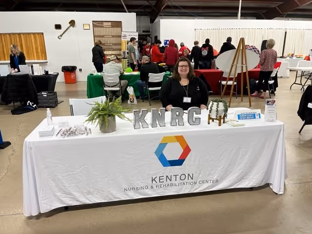 A woman sitting behind a table covered with a white tablecloth displaying the logo and name Kenton Nursing & Rehabilitation Center. The table has decorative plants, letters spelling KNRCC, and informational materials. In the background, several people are standing and interacting at other tables in a large indoor space.