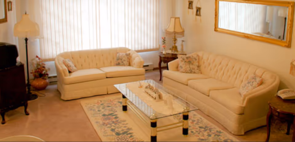 A cozy living room with two cream-colored tufted sofas adorned with floral cushions, a glass coffee table with decorative items on a floral rug, a floor lamp, side tables with lamps and a telephone, a large window with vertical blinds, and a large framed mirror on the wall.
