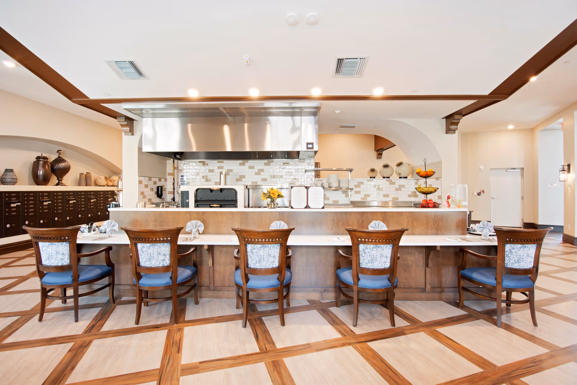 A modern dining area with a long counter and six wooden chairs with blue cushions and patterned backs. Behind the counter is a kitchen area with a stainless steel hood, tiled backsplash, and various kitchen equipment. The floor has a light wood pattern with darker wood inlays, and decorative vases are displayed on shelves to the left.