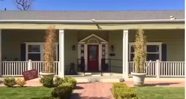 Single-story building front with a covered porch, red entry door, potted plants and a brick walkway through a small manicured lawn.
