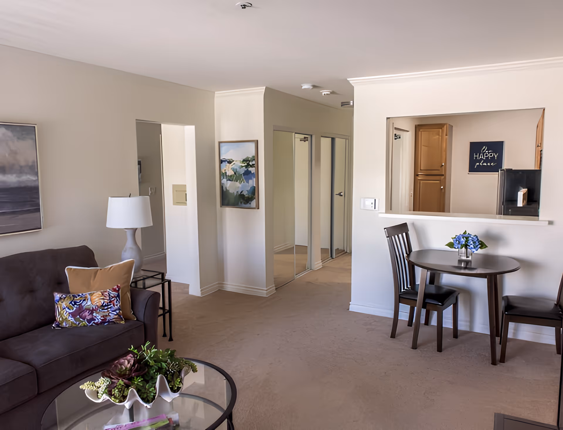 A bright living room and dining area with a sofa, coffee table, small round dining table, and a pass-through window to the kitchen.