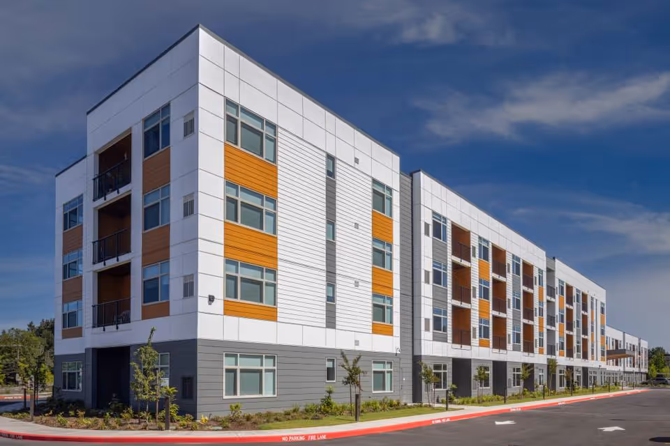 Four-story modern senior living building with white, gray, and orange panels, rows of windows and balconies along a curbside driveway under a blue sky.