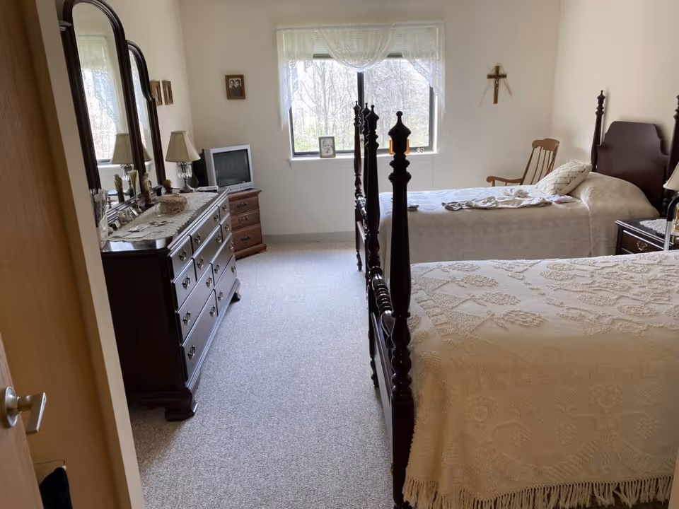 A bedroom with two twin beds covered in cream-colored crocheted bedspreads. There is a wooden dresser with a large mirror on the left side, a small TV on a wooden stand near the window, and a wooden rocking chair in the corner. The window has sheer white curtains, and a cross hangs on the wall above the rocking chair.
