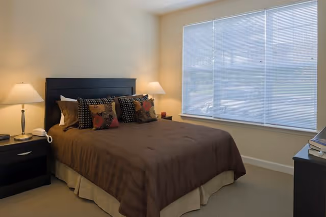 A bedroom with a large bed covered in a brown bedspread and multiple decorative pillows. There are two bedside tables, each with a lamp, and a large window with blinds letting in natural light. The walls are painted a light beige color.