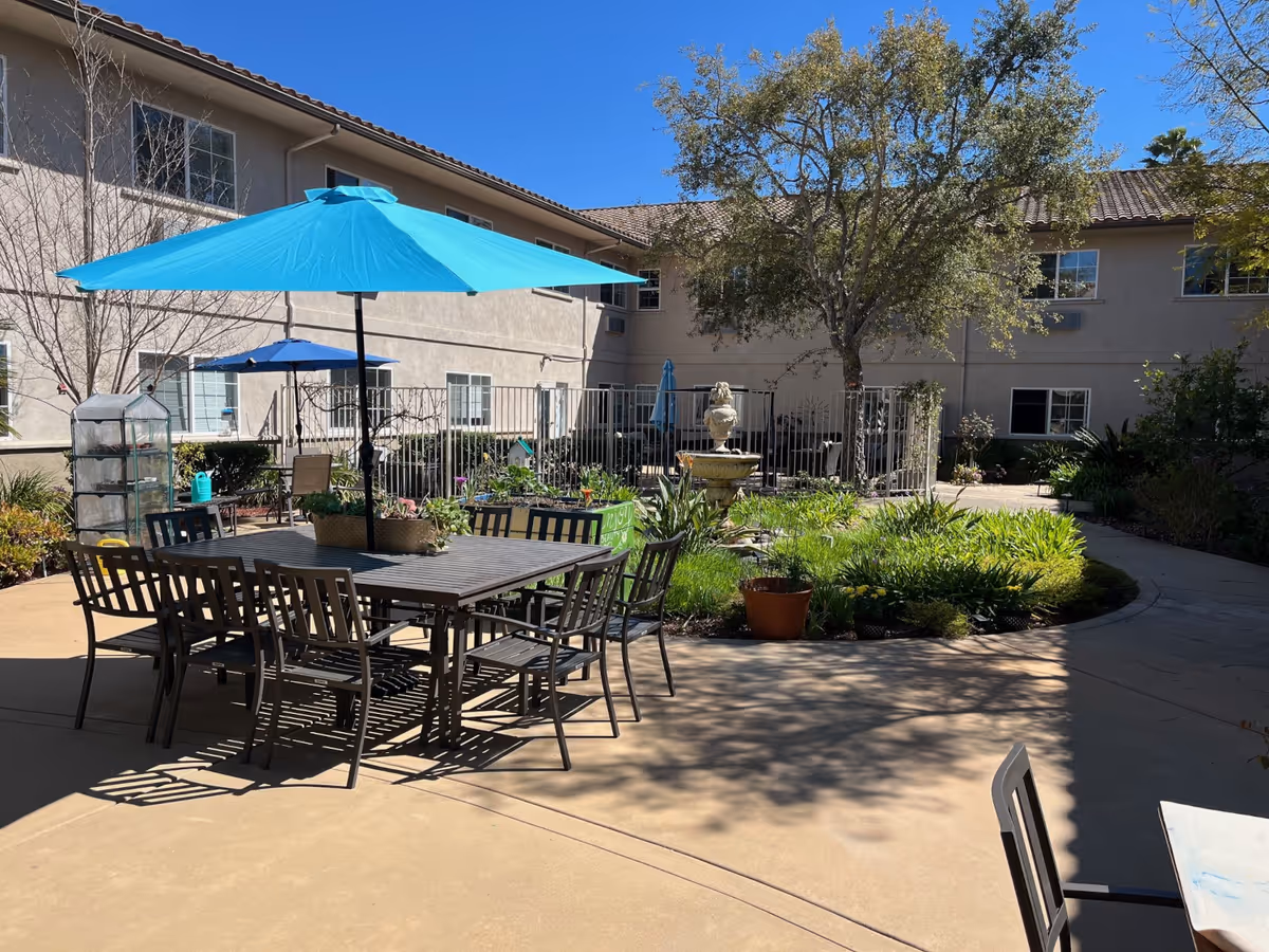 Outdoor courtyard area at Regency Fallbrook with a large table and chairs under a bright blue umbrella, surrounded by plants and trees, with a two-story building in the background under a clear blue sky.