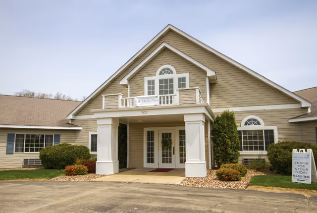 Front exterior view of Oak Creek Place Assisted Living Community building with beige siding, white trim, a covered entrance supported by two columns, and landscaping including bushes and small trees.