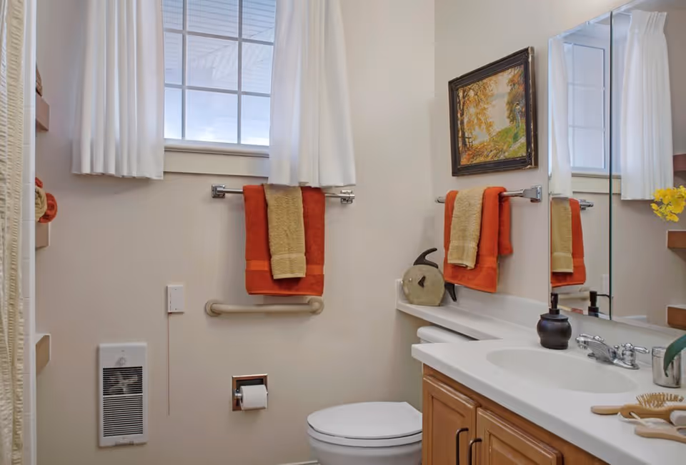 A clean bathroom with a toilet, a white countertop with a sink, a large mirror, and wooden cabinets. There are orange and beige towels hanging on towel racks, a small heater on the wall, a window with white curtains, a framed painting on the wall, and some decorative items on the countertop.