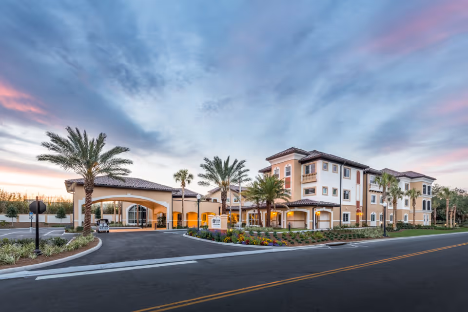 Exterior view of Watercrest Spanish Springs senior living facility at sunset, showing a multi-story building with Mediterranean-style architecture, palm trees, landscaped flower beds, and a covered entrance with a driveway.