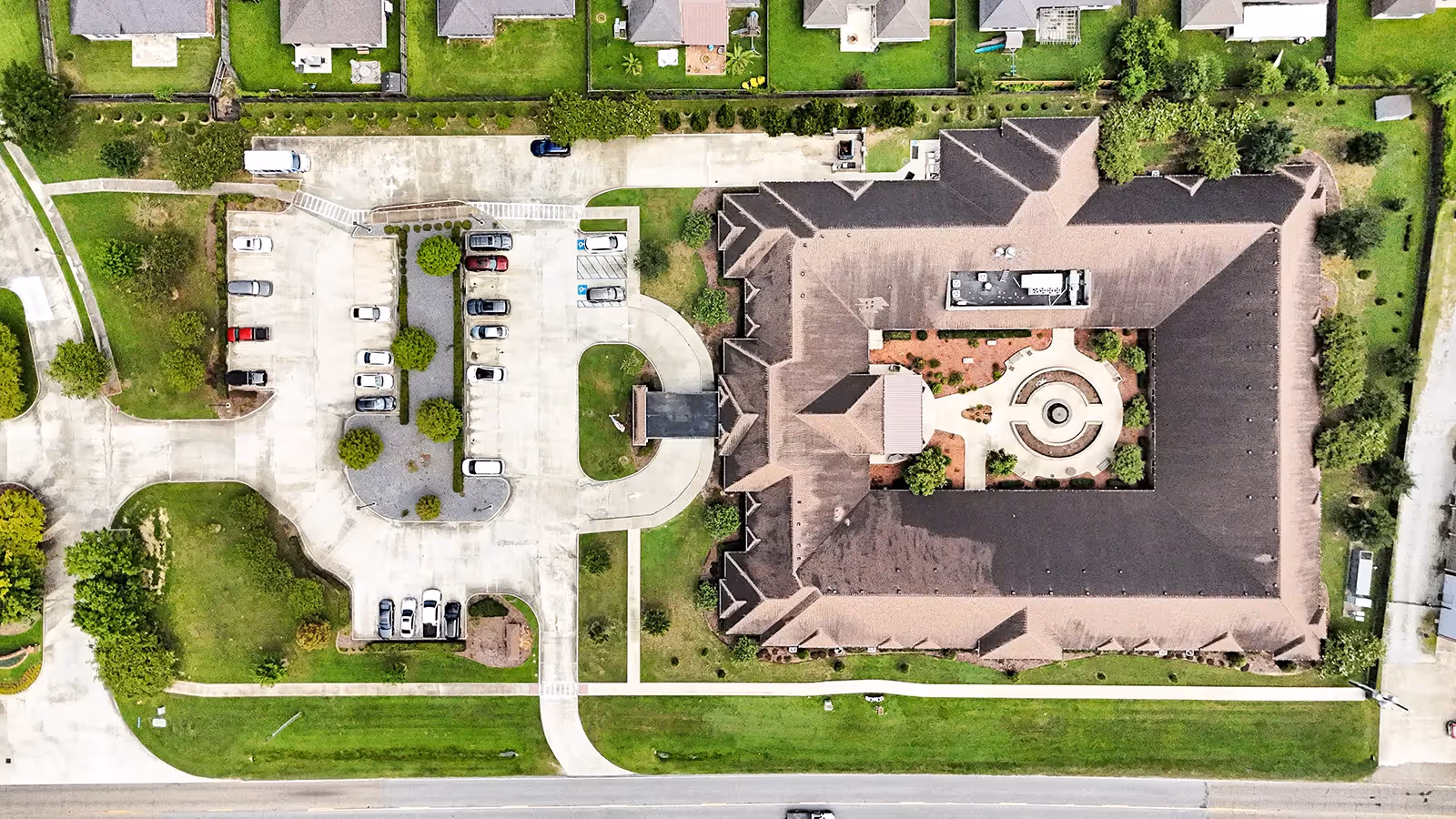 Aerial view of The Pearl at Jamestown - Memory Care Community showing a large building with a central courtyard featuring a circular fountain. Surrounding the building are green lawns, trees, and a parking lot with several cars parked. Residential houses and streets are visible in the background.