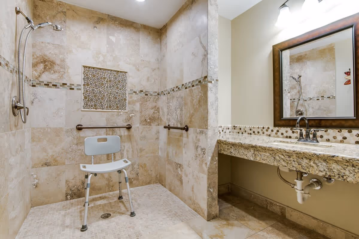 Accessible bathroom with beige tiled walls and floor, featuring a walk-in shower area with a handheld showerhead, a white shower chair, and grab bars. To the right, there is a granite countertop with a sink, a faucet, and a large framed mirror above it.