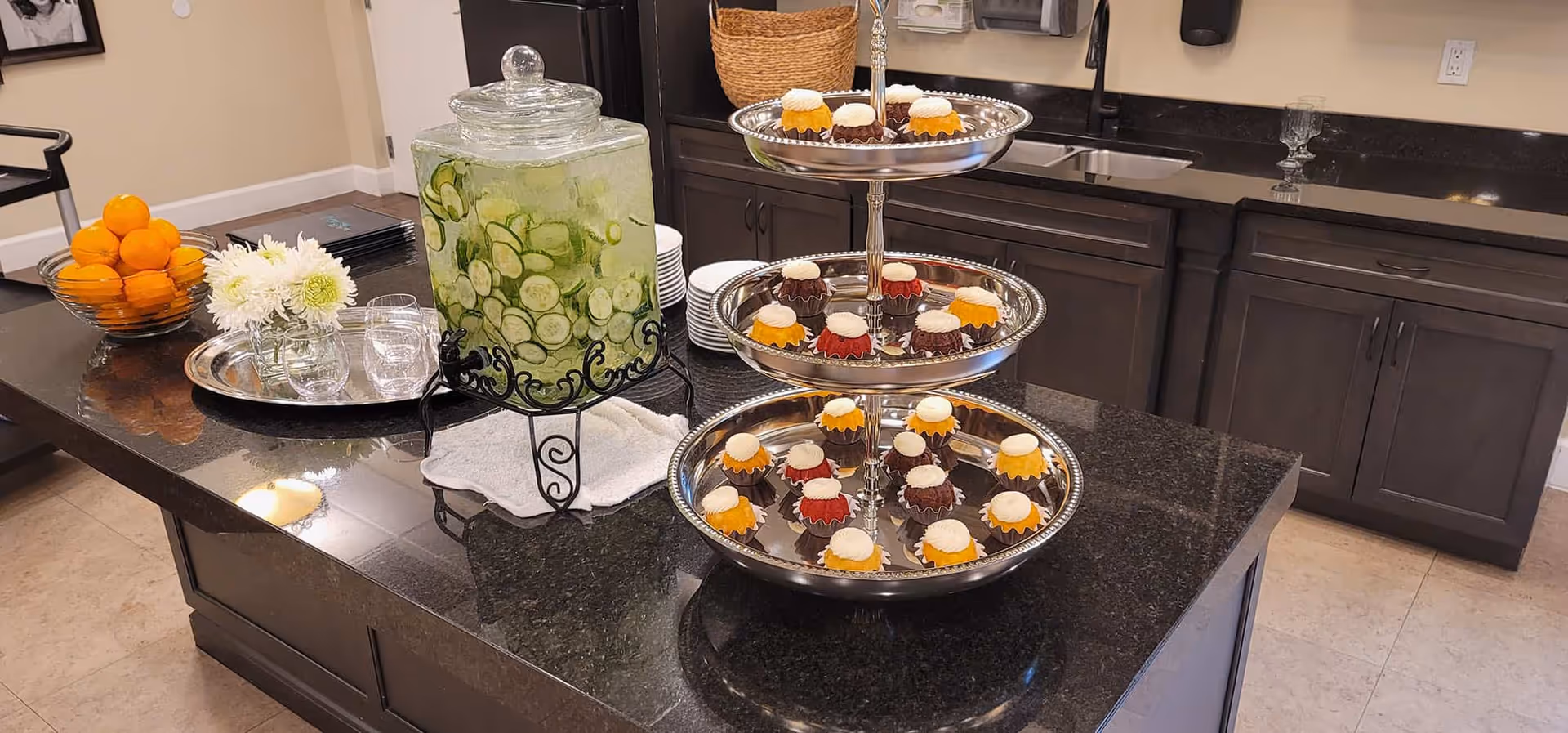 A kitchen island with a three-tiered silver tray holding assorted mini bundt cakes with white frosting. Next to it is a large glass beverage dispenser filled with water and cucumber slices, placed on a white cloth. A silver tray with two glasses and a small bouquet of white flowers in a glass jar is also on the island. In the background, there are dark wood cabinets, a sink, and a bowl of oranges on the counter.