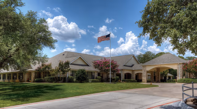 Exterior view of a single-story senior living facility building with a large covered entrance, an American flag on a flagpole in front, surrounded by green lawns, trees, and flowering bushes under a partly cloudy blue sky.
