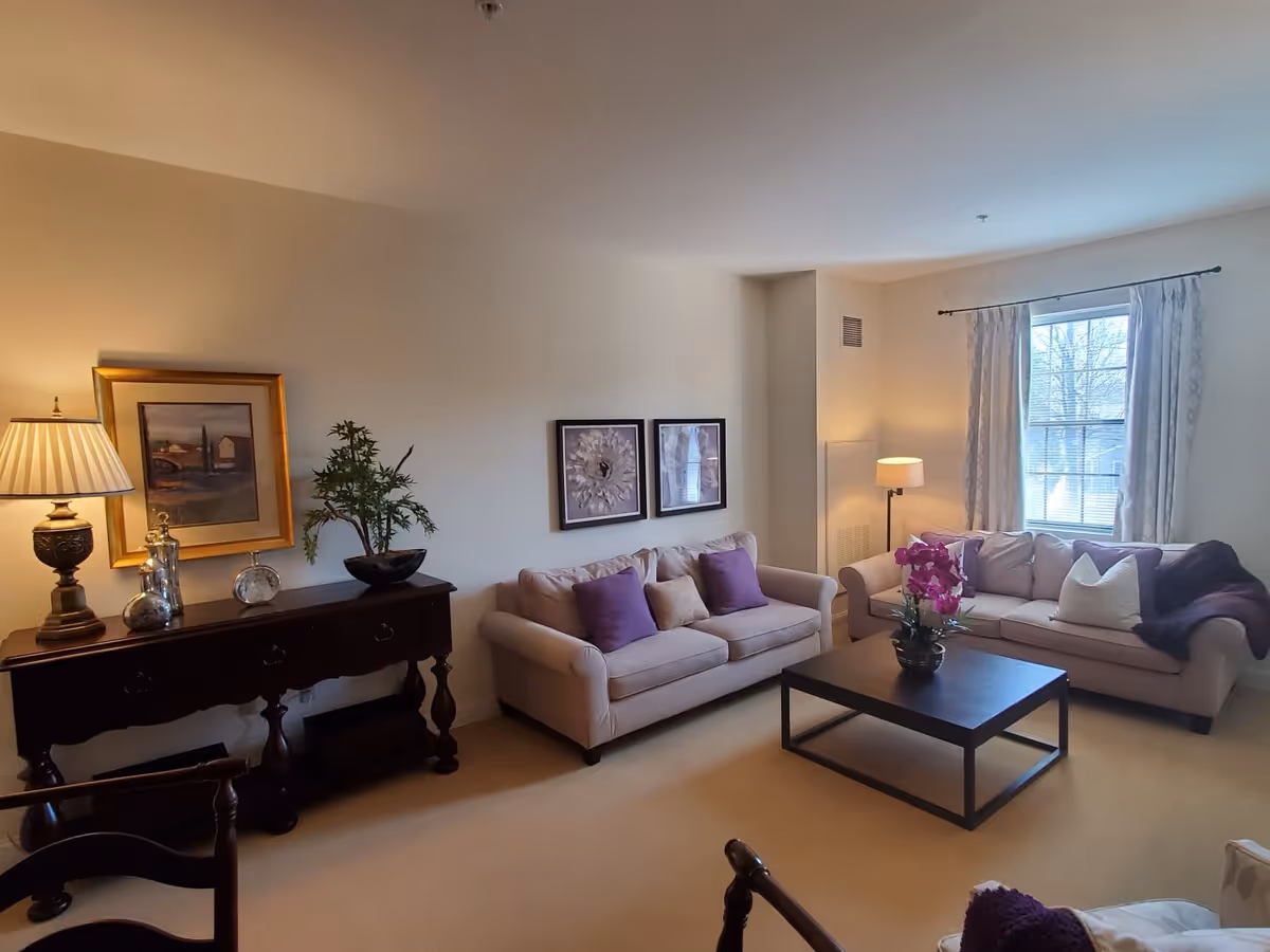A cozy living room with two beige sofas adorned with purple and white pillows, a dark wooden coffee table with a purple orchid centerpiece, a side table with a lamp, framed artwork on the walls, and a window with light curtains letting in natural light.