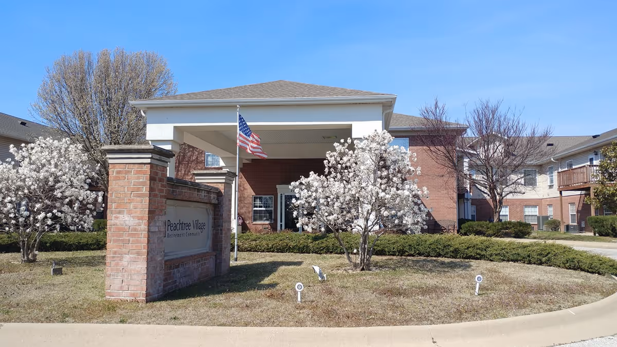 Front exterior of Peachtree Village senior living building showing the covered entrance, brick sign, American flag, and flowering trees.