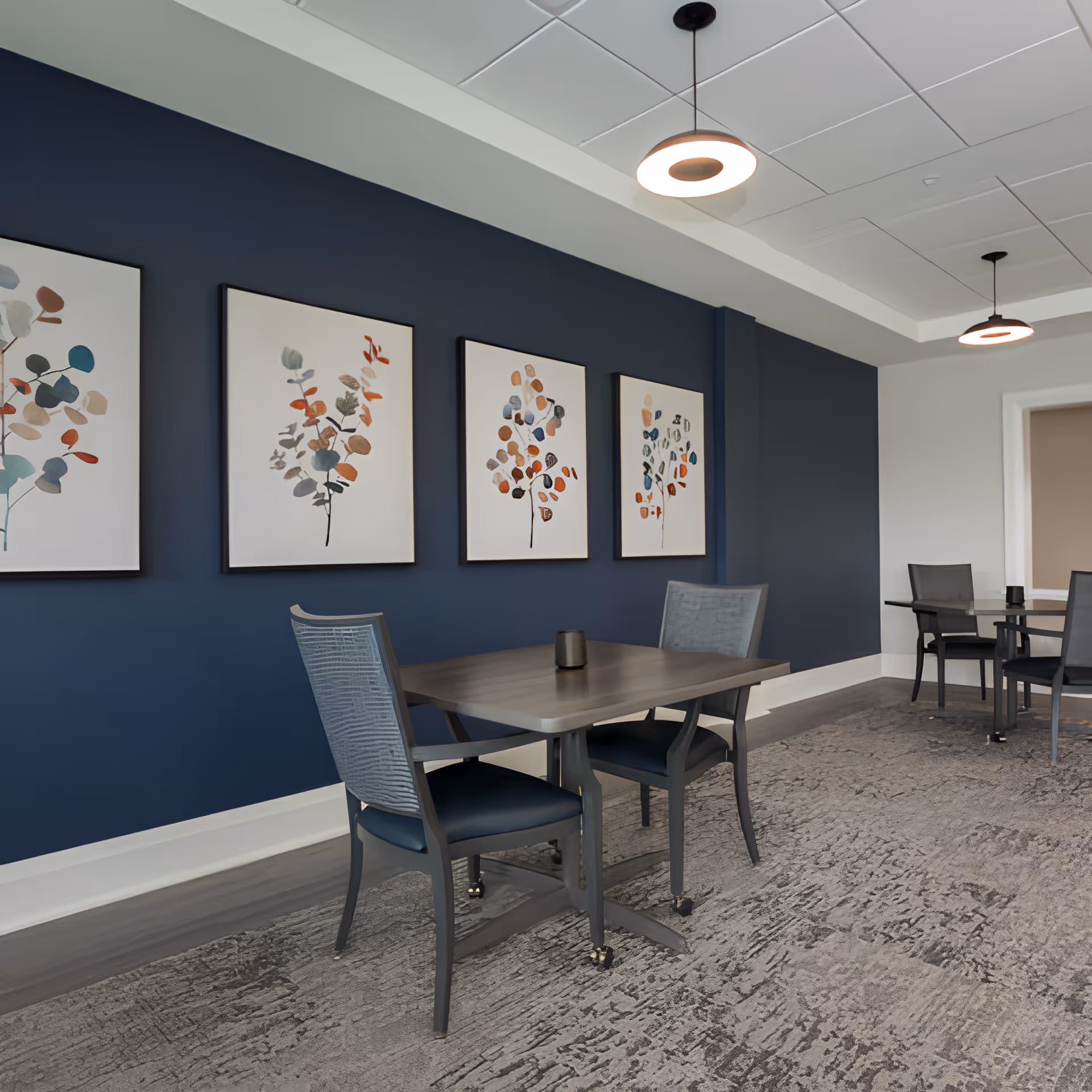 Small dining/activity room with tables and chairs, a navy accent wall featuring four framed botanical prints, and pendant lights.