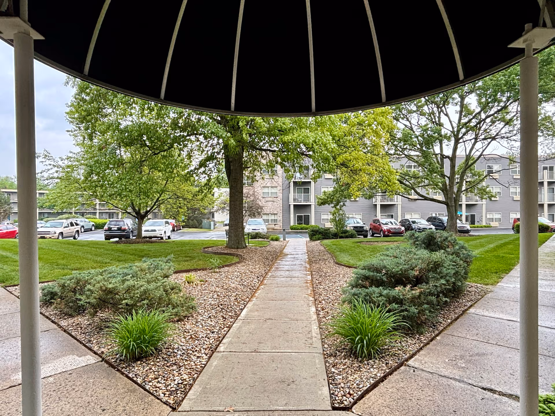View from under a black canopy looking out onto a landscaped pathway with green bushes and trees on either side, leading to a parking lot with several cars and a multi-story residential building in the background.