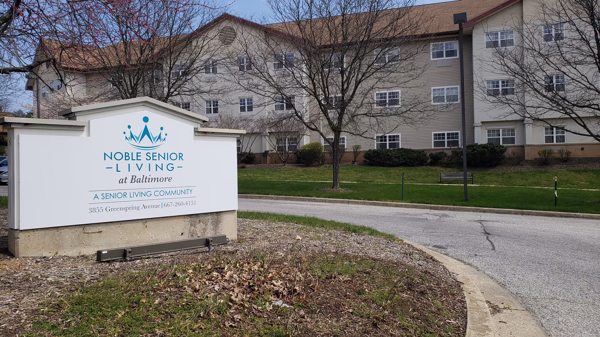 Outdoor view of the entrance to Noble Senior Living at Baltimore, showing a large white sign with the facility's name, logo, and contact information. In the background, there is a multi-story building with multiple windows, leafless trees, and a curved driveway.