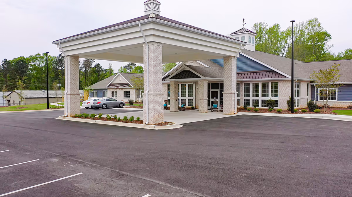 Exterior view of The Colonial at Old Camden senior living facility showing the main entrance with a covered drop-off area, parking lot, and surrounding greenery.