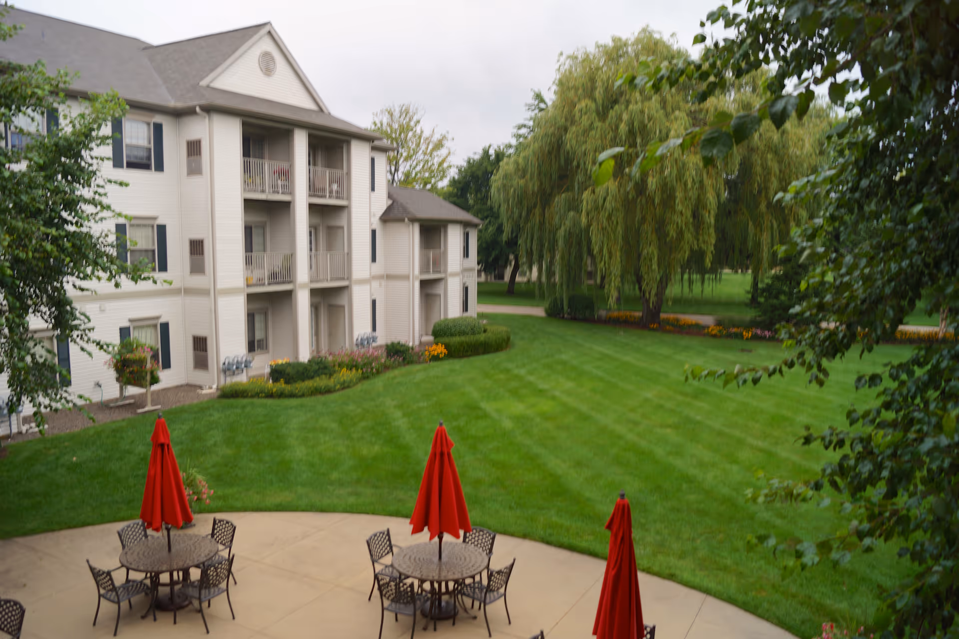 Outdoor patio area with round tables and red umbrellas, surrounded by green grass and trees, adjacent to a multi-story residential building with balconies.