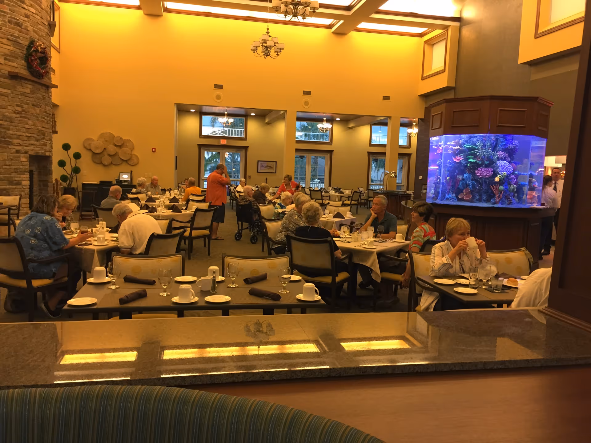Residents dining in a large, warmly lit communal dining room with an aquarium and high ceilings.