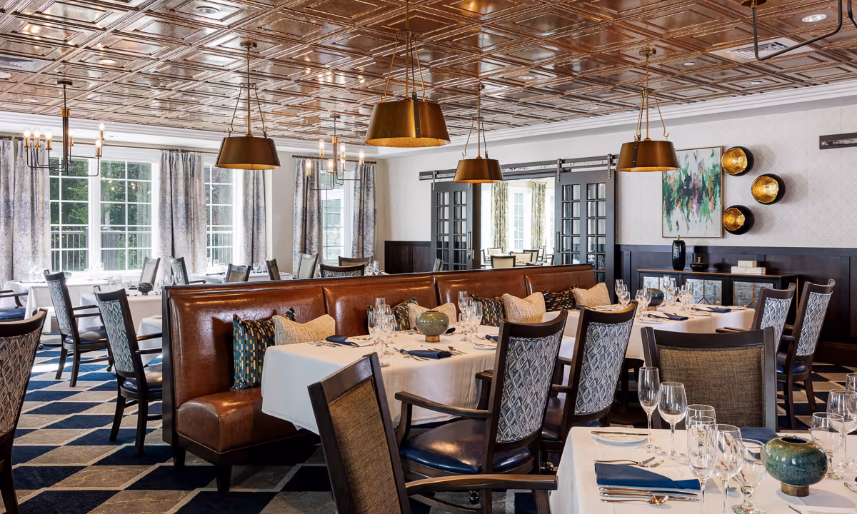 Elegant dining room with tables set with white tablecloths, glassware, and napkins. The room features a decorative ceiling, pendant lighting, patterned chairs, and a leather bench with cushions. Large windows with curtains allow natural light to fill the space.